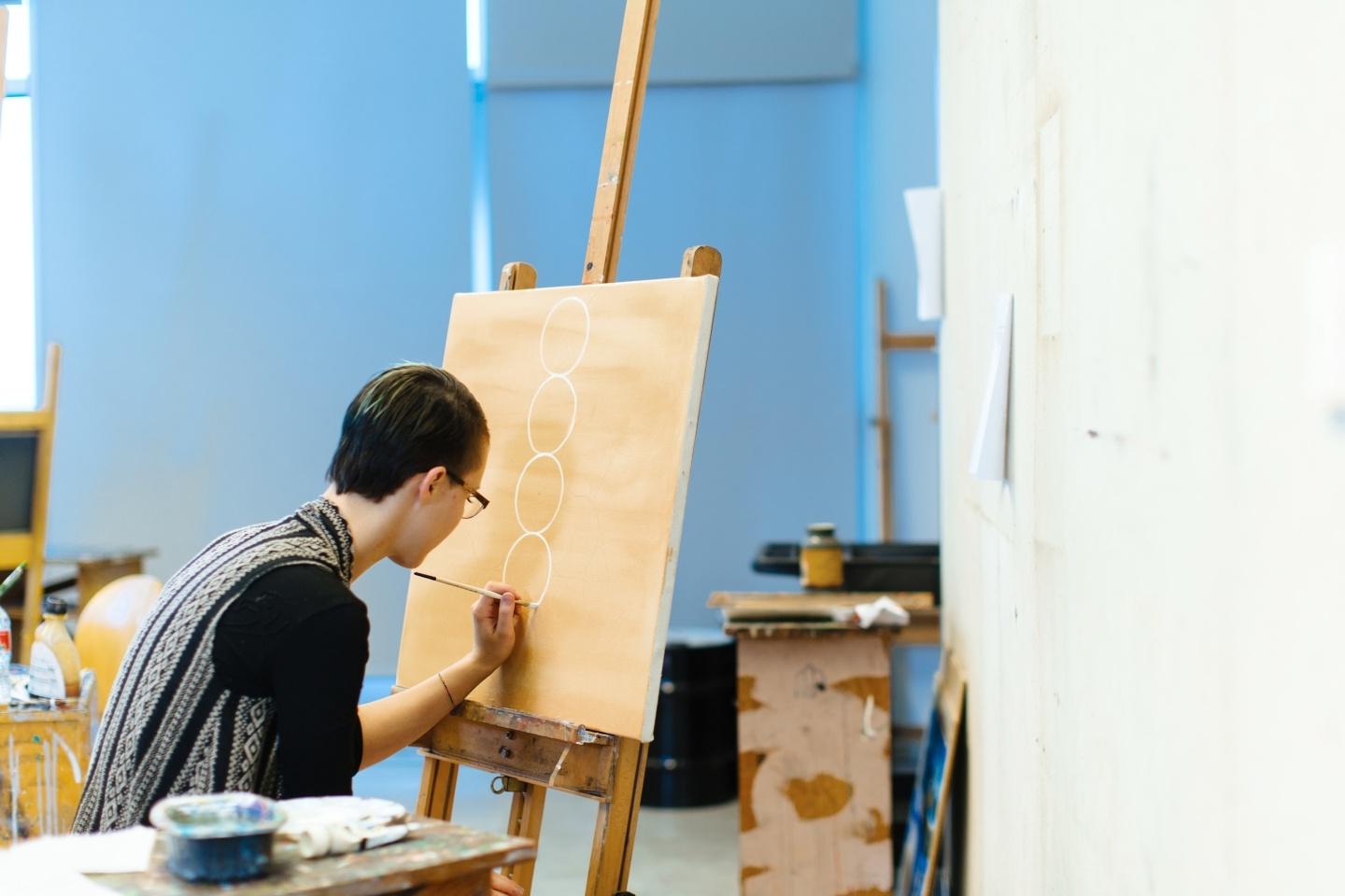 A student paints circles on a canvas in a naturally-lit art studio
